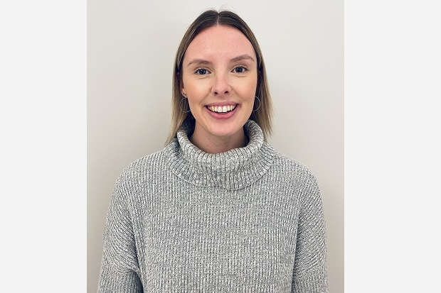 A lady with long, straight, fair hair, wearing a grey polo neck sweater and smiling at the camera, in front of a grey background.