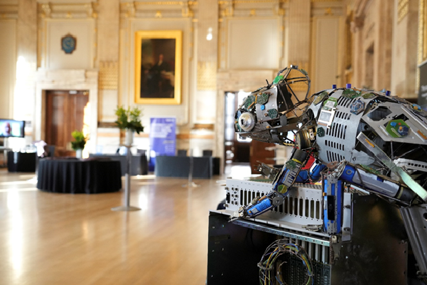 Photograph of a robotic sculpture resembling a crouching animal, displayed in an ornate room with wooden floors and classical decor. Sculpture features exposed mechanical parts, wires, and metallic panels, highlighting technological artistry within a formal exhibition setting.