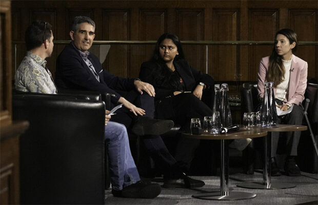 Four speakers seated on a stage during a panel discussion, with microphones and water on small tables between them. The speakers are engaged in conversation in a wood‑panelled hall, as part of a formal conference session.