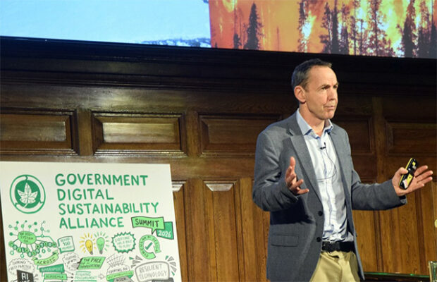 Photograph of a speaker presenting at the Government Digital Sustainability Alliance Summit 2026, with a wooden panelled background and a large illustrated poster highlighting sustainability themes such as renewable energy and environmental protection. The speaker gestures with hands while holding a remote, and a projected image of a wildfire contrasts with a glacier, emphasizing climate change challenges.