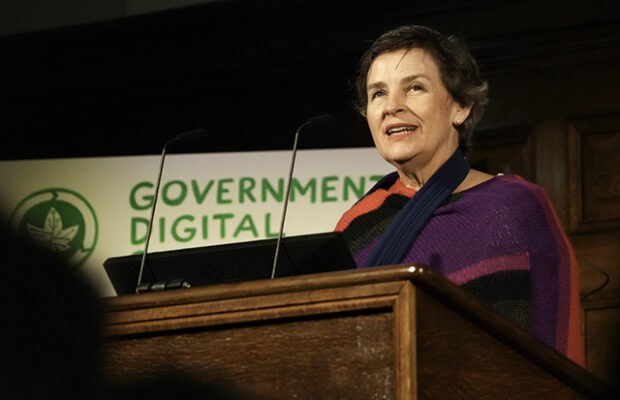 Photograph of a person standing behind a wooden podium with microphones, delivering a speech at an event. A sign behind the podium reads "Government Digital," indicating a conference or awards ceremony related to digital government services.