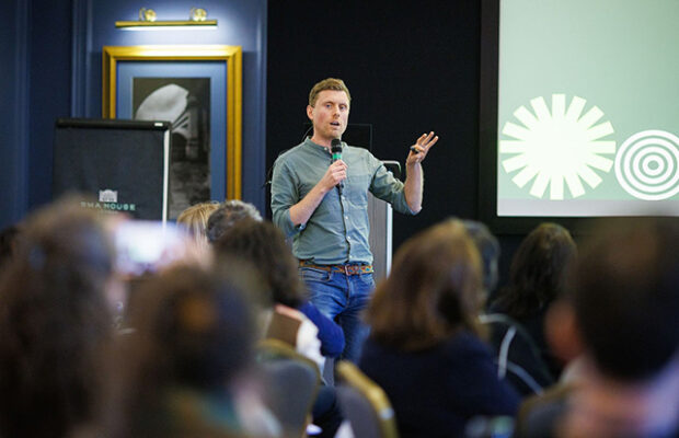 A speaker standing at the front of a room, holding a microphone and gesturing while presenting to a seated audience. A presentation slide is displayed behind the speaker, and the audience is visible in the foreground.