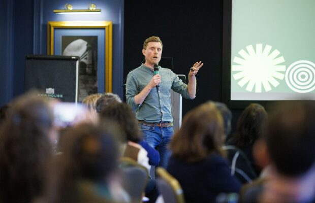 A speaker standing at the front of a room, holding a microphone and gesturing while presenting to a seated audience. A presentation slide is displayed behind the speaker, and the audience is visible in the foreground.