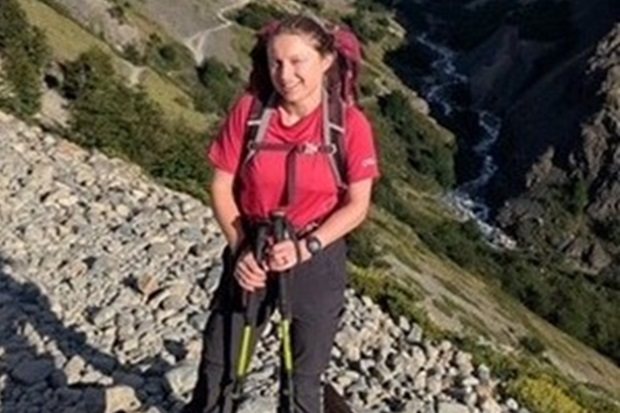 A girl with a red t shirt, grey trousers and walking poles. With a valley and a stream in the background.