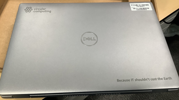 Photograph of a closed silver Dell laptop with "circular computing" logo in top left corner and slogan "Because IT shouldn't cost the Earth" in bottom right corner. Laptop is placed on a wooden surface with a barcode sticker on top right corner.